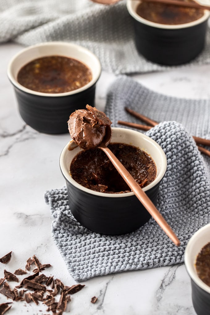 A bronze spoon resting on a black pot filled with chocolate creme brulee. Some chopped chocolate in front