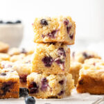 A stack of 3 lemon blueberry bars, surrounded by more and fresh blueberries.