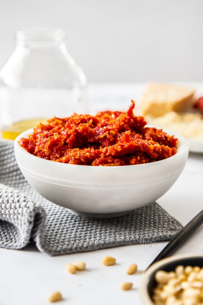 A white bowl filled with tomato pesto, surrounded by the ingredients that go into it. It sits on a grey napkin