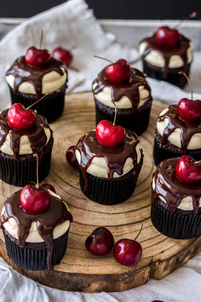 A batch of chocolate and cherry topped cupcakes on a wooden board.