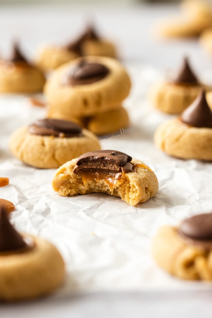 A Peanut butter thumbprint cookie with a bite taken out of it, showing caramel in the middle. It sits on a sheet of baking paper with others around.