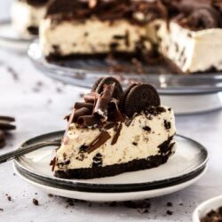 A slice of no bake oreo cheesecake sitting on a white plate on a concrete table. Oreos and crumbs strewn around it