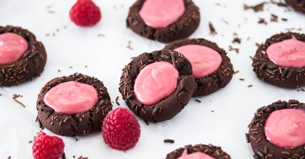 Some Chocolate Raspberry Thumbprint Cookies and fresh raspberries scattered around on a white surface.