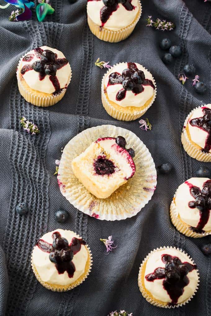 A Lemon Blueberry Cupcake cut in half showing the blueberry compote centre. More cupcakes and blueberries are scattered around.