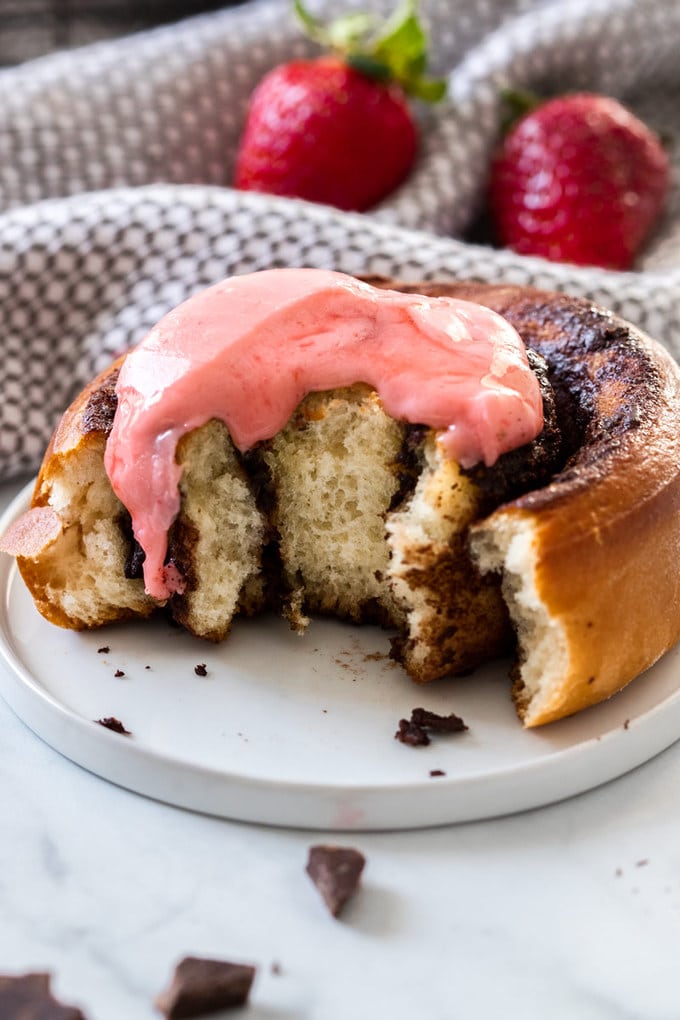 A chocolate cinnamon roll with pink icing on a white plate, with a bite taken out