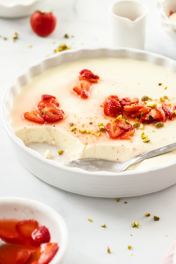 A large white dish filled with panna cotta. A spoon sits where some has been taken and some macerated strawberries sit on top and in front.
