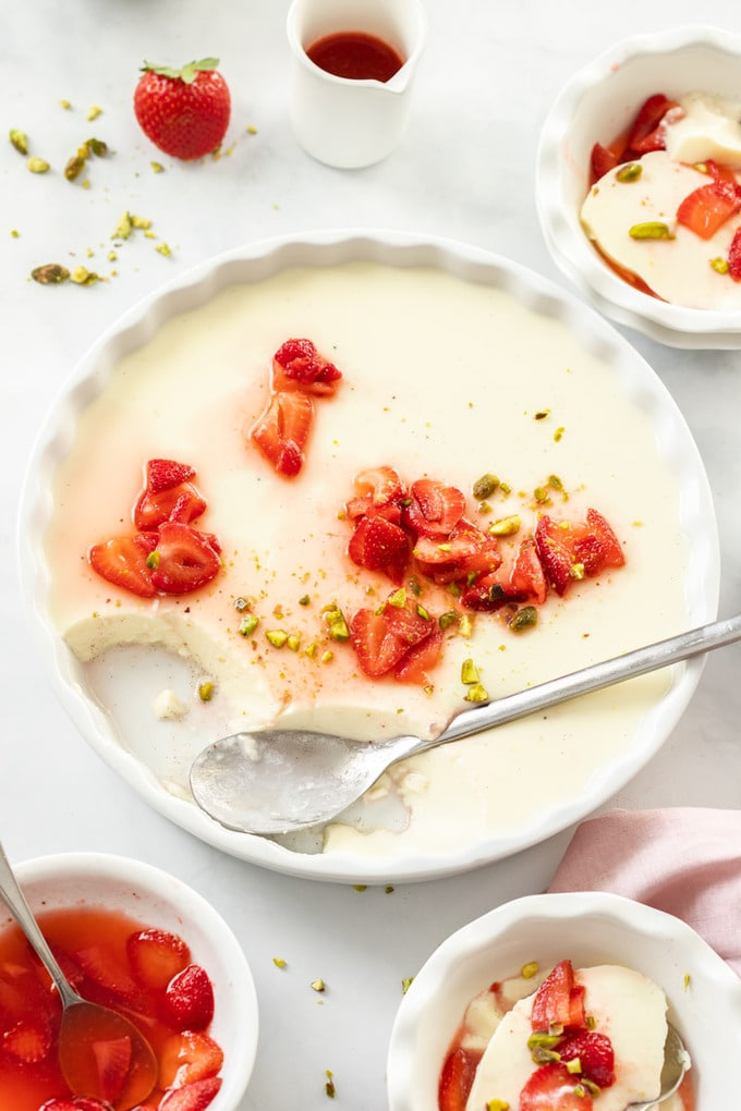 A large white dish filled with panna cotta. A spoon sits where some has been taken and some macerated strawberries sit on top and in front.