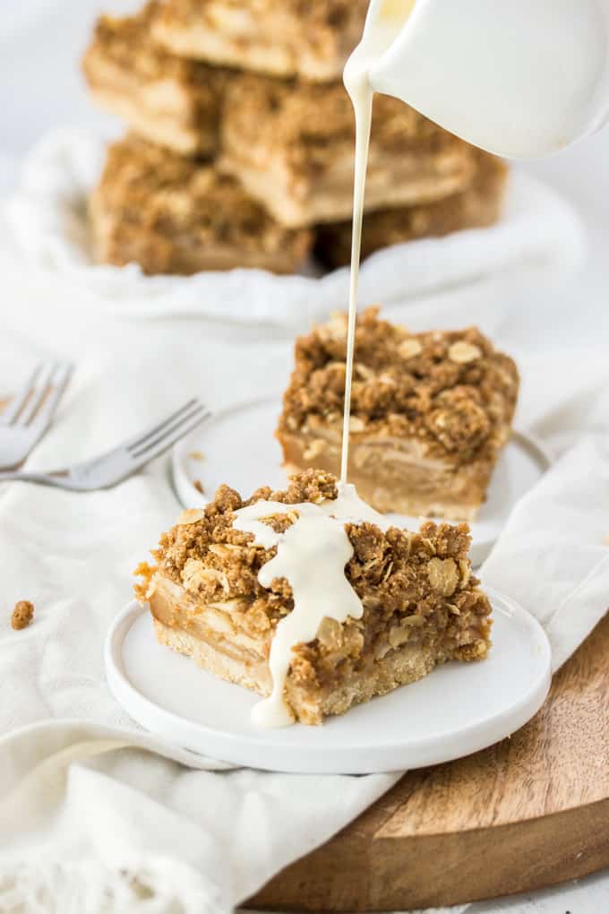 Cream being poured over a caramel apple pie bar on a white plate with more slices in the background.