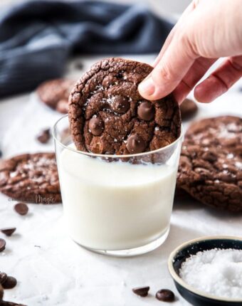 A chocolate cookie being dunked into a glass of milk with more cookies scattered around it