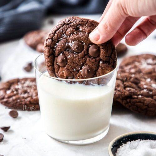 A chocolate cookie being dunked into a glass of milk with more cookies scattered around it