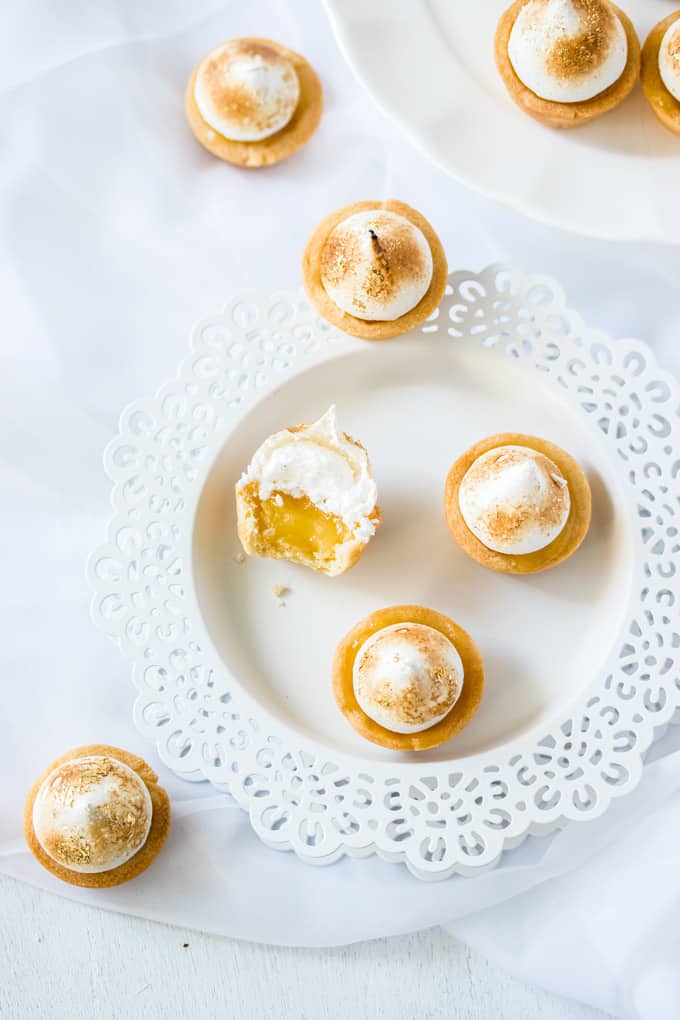 3 Mini Lemon Meringue Pies on a plate, one is half eaten, showing the inside. More mini pies scattered around.