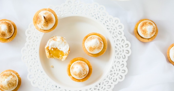 Mini Lemon Meringue Pies on a white plate, one is half eaten, showing the filling inside.