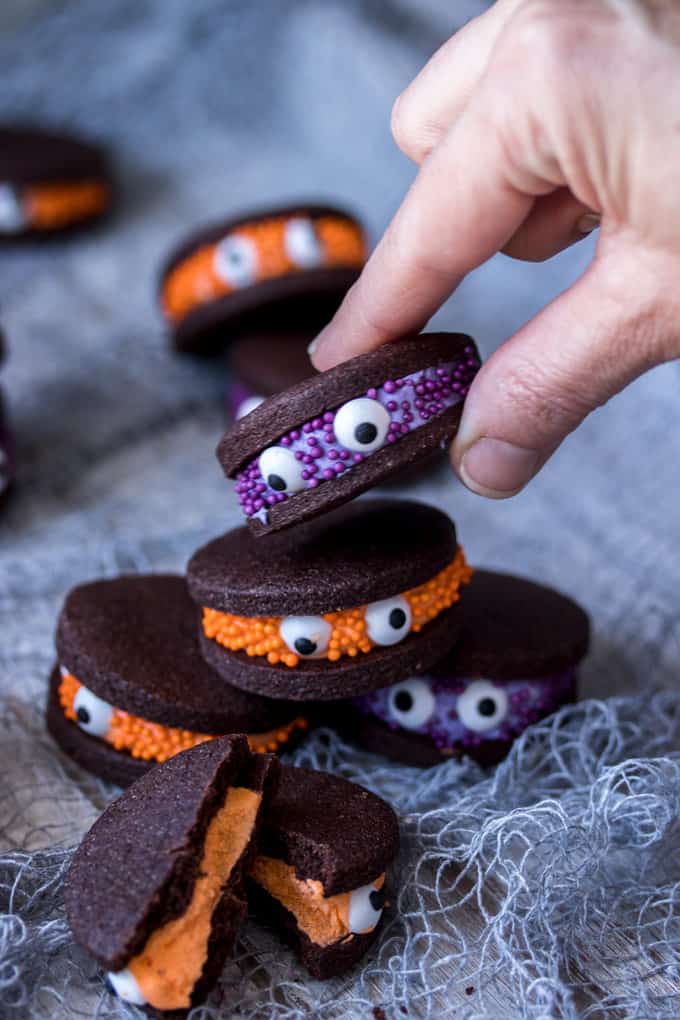 A hand picking up a chocolate sandwich cookies with purple filling, decorated with purple sprinkles and eye balls.