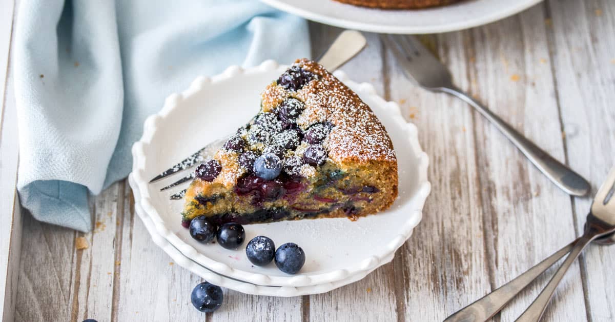 A slice of Blueberry Cake on a white plate with some fresh blueberries.