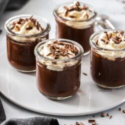 4 little glasses filled with chocolate pudding on a white plate with chocolate curls around