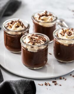 4 little glasses filled with chocolate pudding on a white plate with chocolate curls around