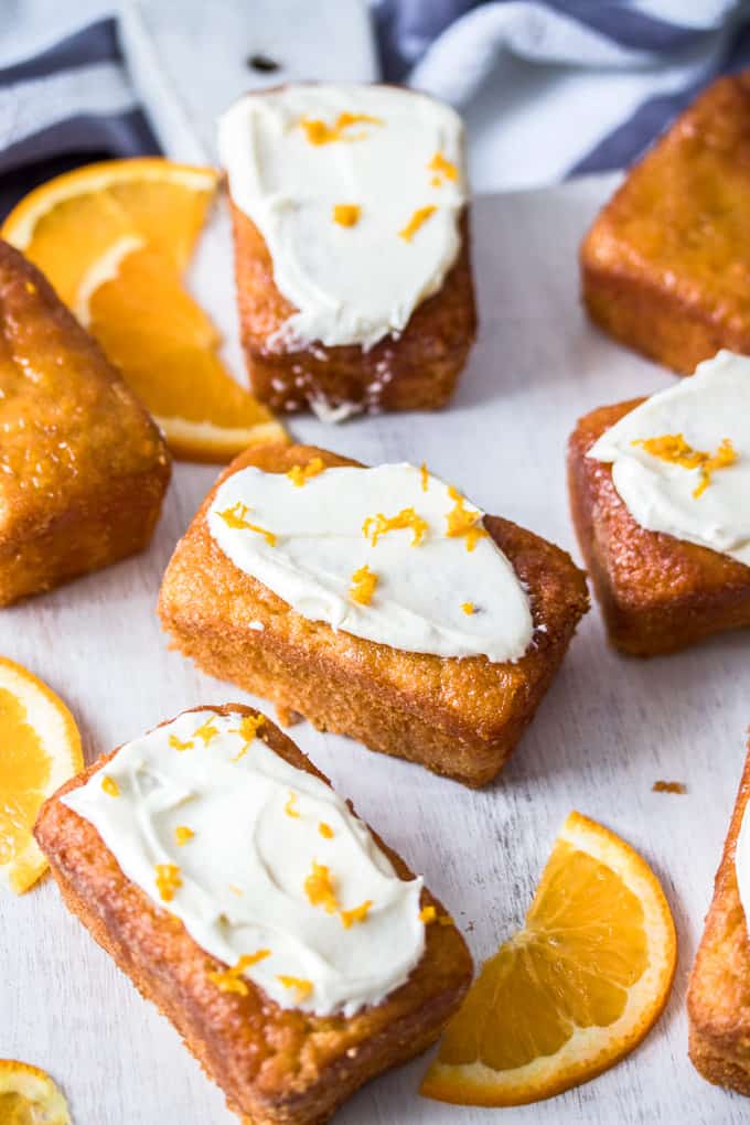 A few Mini Flourless Ginger Orange Cakes, some with white frosting, on a white board, next to orange slices.