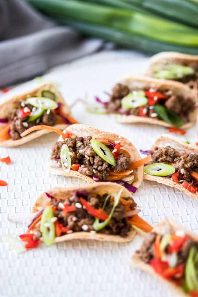 A group of mini tacos filled with ground beef bulgogi and Asian slaw on a white mat with green onions in the background.