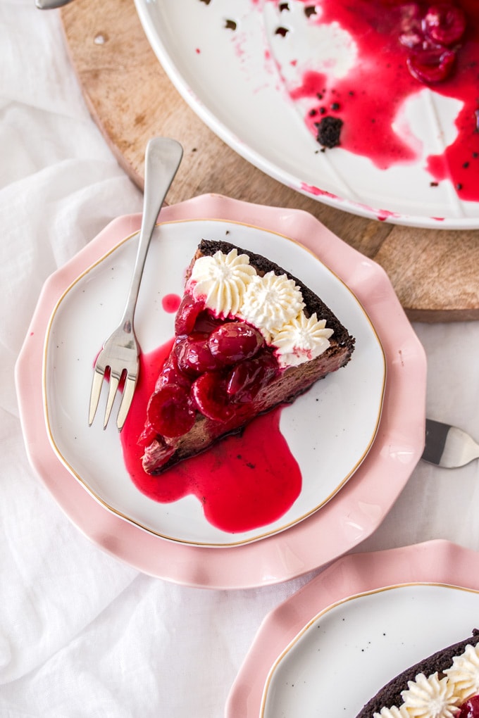 A slice of chocolate cheesecake topped with cream and cherry sauce on a white cake plate with a fork next to it.