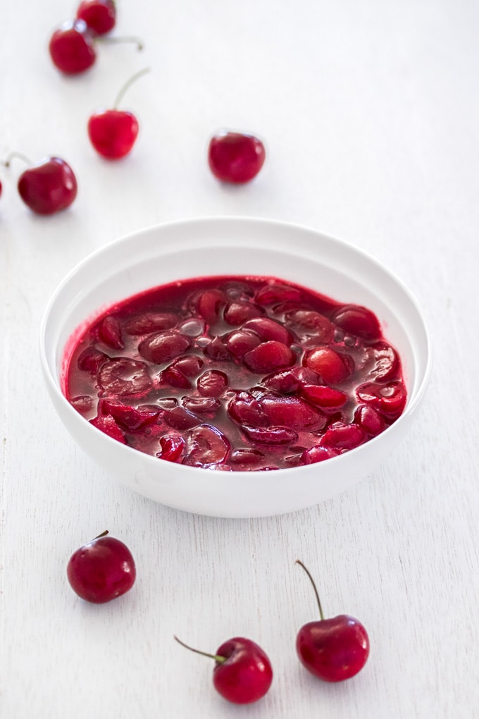 A white bowl filled with cherry sauce on a white wooden background with fresh cherries around it.