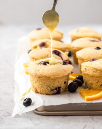 Muffins sitting on a sheet of baking paper on a baking tray. surrounded by blueberries and slices of orange.