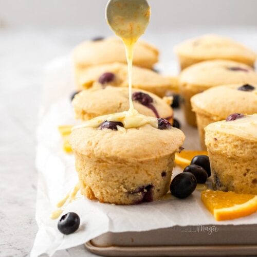 Muffins sitting on a sheet of baking paper on a baking tray. surrounded by blueberries and slices of orange.