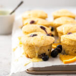 Muffins sitting on a sheet of baking paper on a baking tray. surrounded by blueberries and slices of orange
