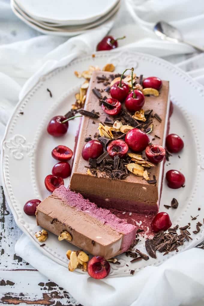 A pink and brown frozen parfait on a plate covered in cherries and chocolate