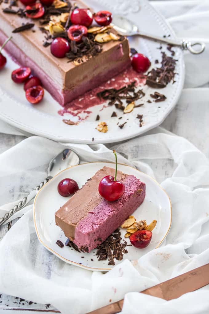 A slice of pink and brown parfait on a white plate with a cherry on top