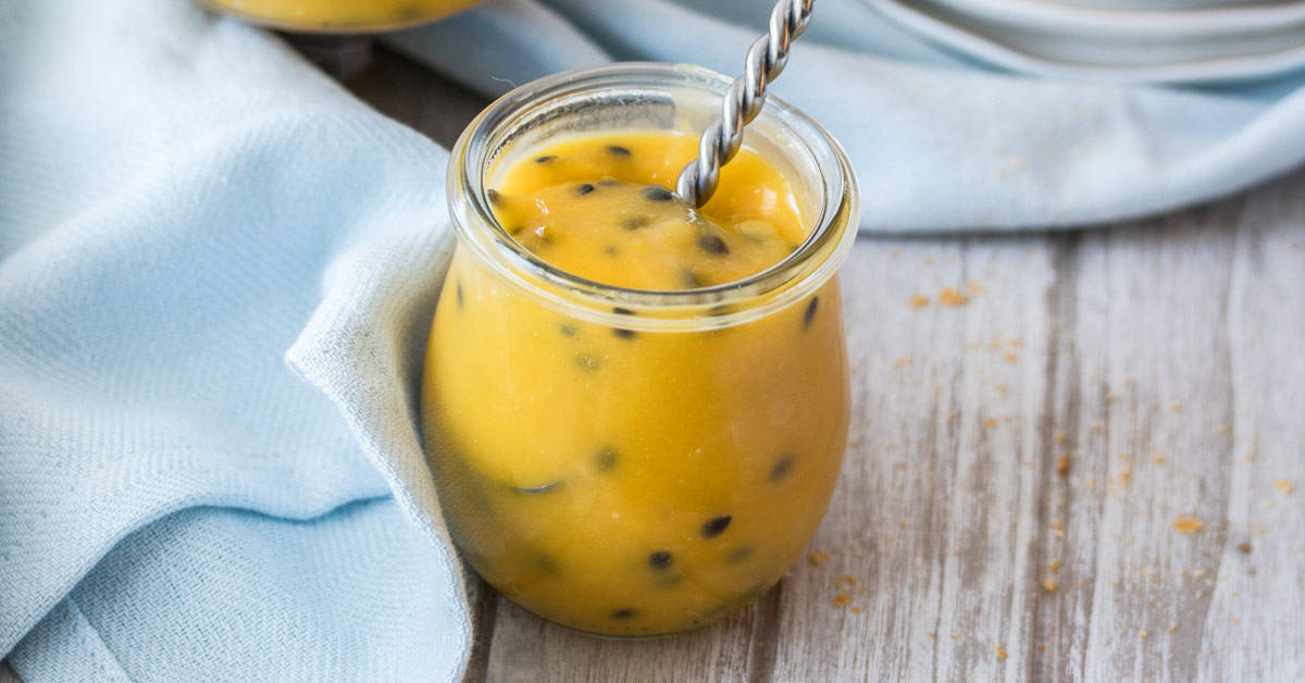 Passionfruit Curd in a glass jar with a small spoon handle sticking out, next to a blue towel.
