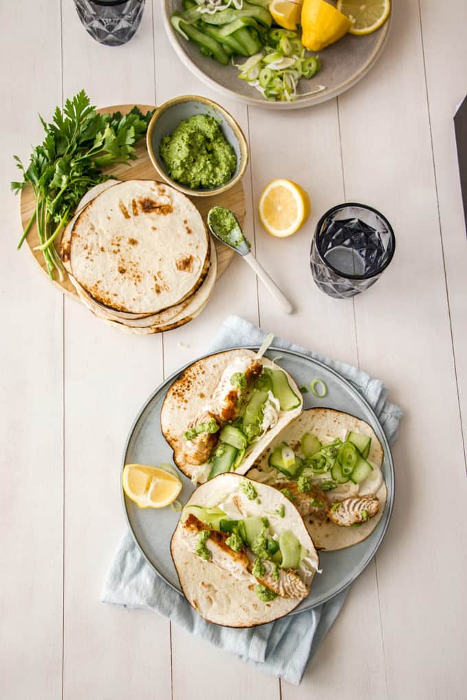 Overhead view of Fish Tacos on a large plate on top of a blue towel, a round board with tortillas, pesto and garnishing