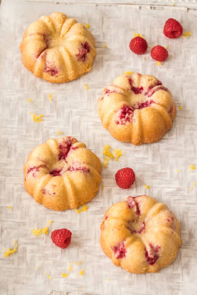 4 Lemon Raspberry Mini Bundt Cakes sitting on a textured surface with raspberries and lemon zest sprinkled about