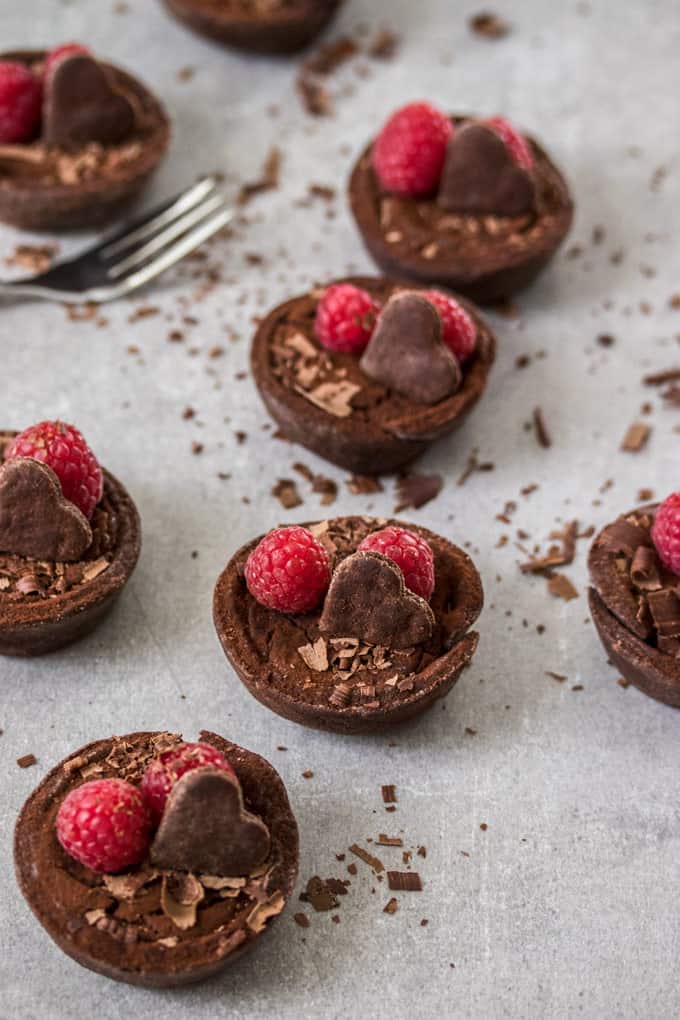 4 mini chocolate tarts lined up, decorated with heart shaped cookie and fresh raspberries