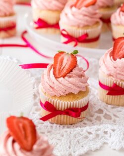 Six Vanilla cupcakes with strawberry buttercream with strawberries on top.