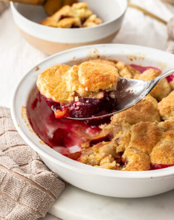 A spoon filled with cobbler dessert, scooping from a white pie dish.