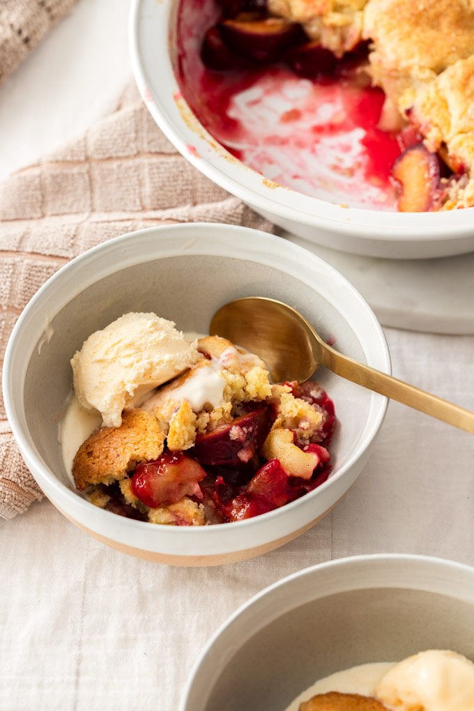 A white bowl filled with cobbler and a scoop of ice cream. A gold spoon on the side.