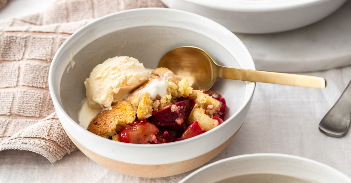 A white bowl filled with cobbler and a scoop of ice cream. A gold spoon on the side.