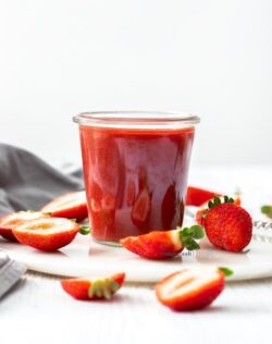 A small glass pot filled with strawberry sauce, surrounded by cut strawberries