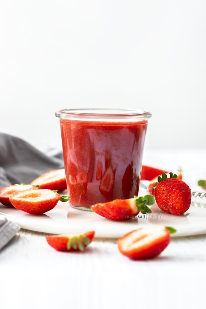 A small glass pot filled with strawberry sauce, surrounded by cut strawberries