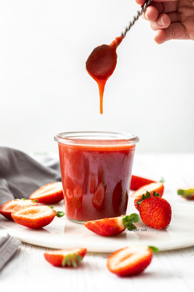 A spoon is lifted from a jar of strawberry sauce, surrounded by cut strawberries