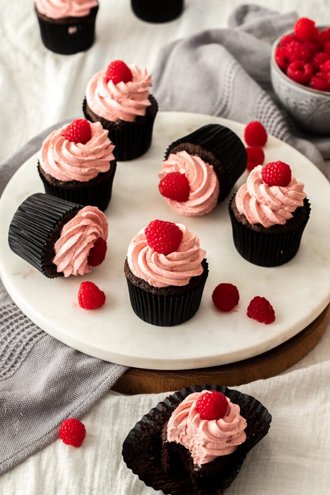 Six chocolate cupcakes with pink frosting on a white platter with fresh raspberries scattered around.