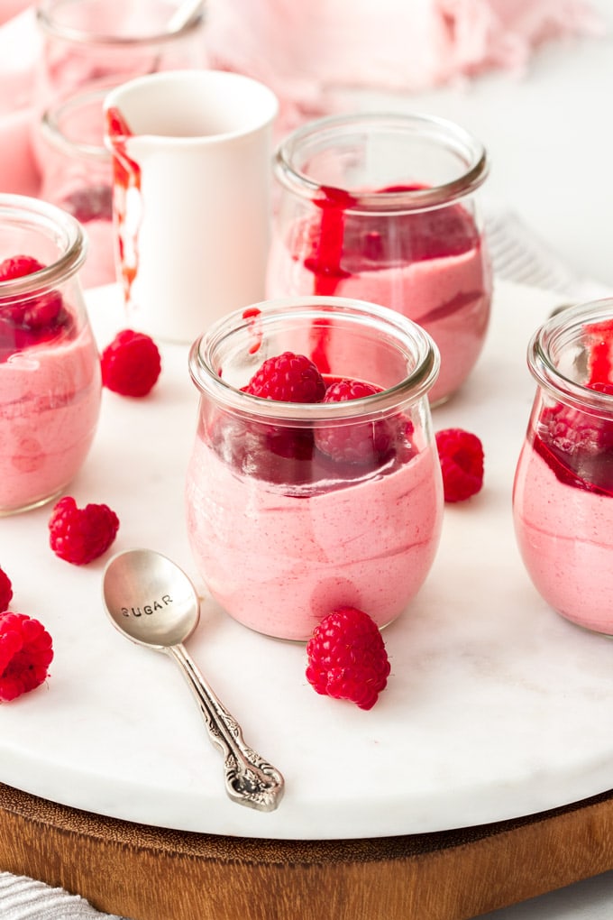A white platter holding 4 small glass jars with raspberry mousse. Fresh raspberries scattered around