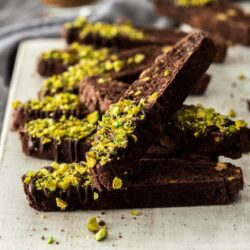 A number of pieces of chocolate biscottion sprinkled with pistachios, piled up on a white board