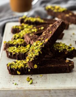 A number of pieces of chocolate biscottion sprinkled with pistachios, piled up on a white board