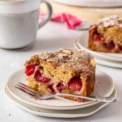 A slice of rhubarb cake sitting on a white plate with a fork next to it, a cup of coffee in the back
