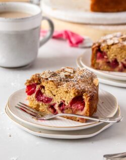 A slice of rhubarb cake sitting on a white plate with a fork next to it, a cup of coffee in the back