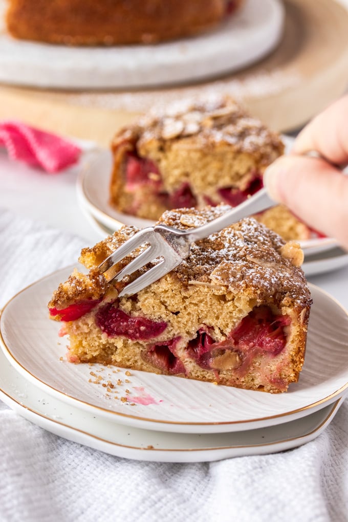 A fork digging into a piece of rhubarb cake on a white plate with gold rim.
