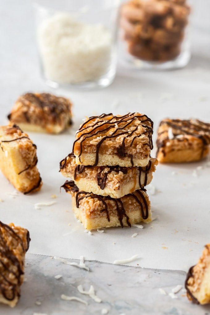 A stack of 3 caramel coconut bars on a piece of baking paper with a glass of coconut in the background
