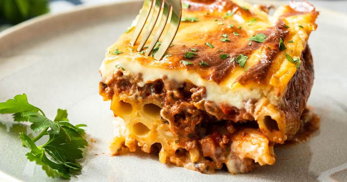 A slice of pastitsio on a grey plate with a fork digging into it
