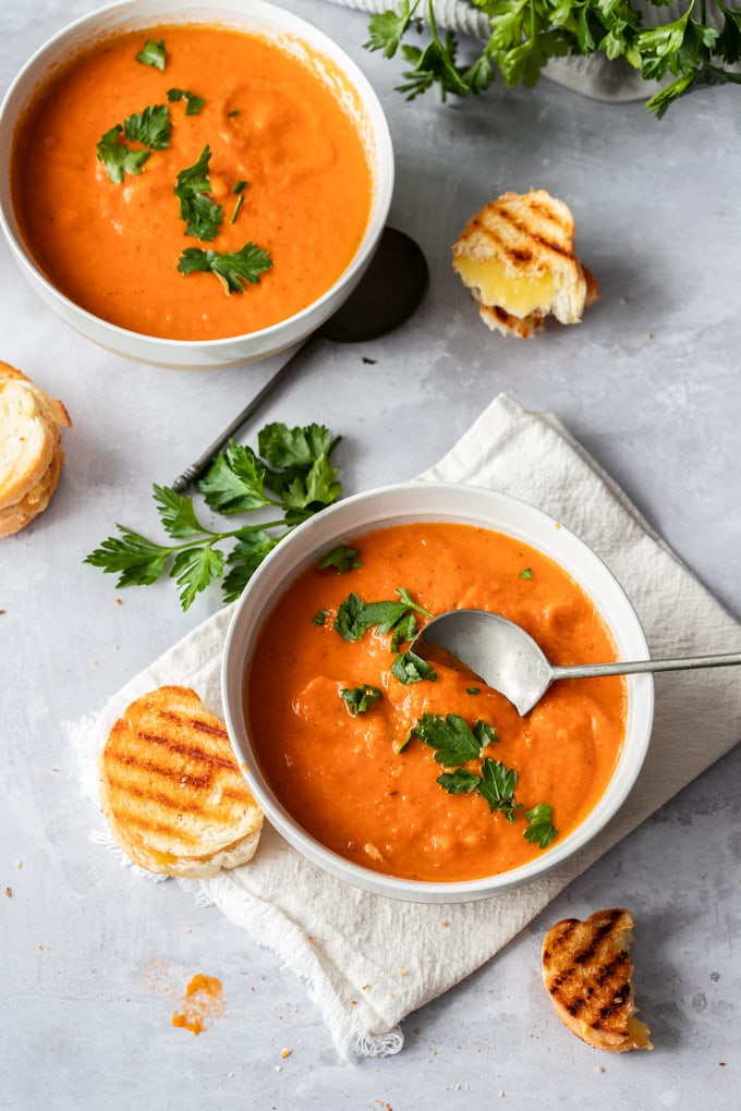 Overhead shot of two bowls of tomato soup with parsley and grilled cheese sandwiches nearby.